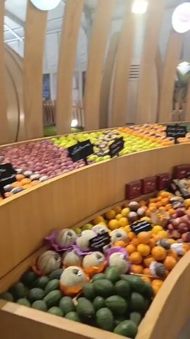 The video captures an indoor market or exhibition space, likely a food fair or fresh produce section. Various fresh fruits and vegetables are artfully displayed on tiered, curved wooden shelves. These include abundant piles of green avocados, bright orange mandarins or oranges, red apples, green apples, and yellow apples, along with other colorful produce. Some packaged goods, possibly chocolates or other snacks, are also visible on the shelves. A large digital display screen is prominent in the background, showing text and a graphic featuring a checkmark within a circular seal. Wooden stools and barrel-shaped tables are placed in the area, suggesting a potential tasting or seating arrangement. The space is brightly lit and features a tent-like ceiling structure. No people are visible, but the setup implies an active market environment in Al Machouar – Stinia, Morocco. Small black labels are present on the produce displays, but their text is not legible.