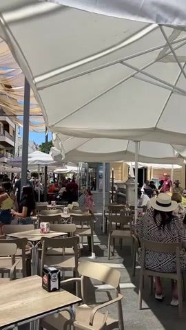 The video shows an outdoor cafe scene on a sunny day. In the foreground, several tables and chairs are set up under large white umbrellas and fabric canopies. A person wearing a patterned top and a hat is seated at one of the tables. In the background, a street is lined with buildings. People are walking along the street, some are seated at outdoor tables of other cafes, and there is a small group of people gathered around two musicians, one playing a guitar and the other a wind instrument. A banner hangs from the building behind the musicians. The overall atmosphere appears lively and social.