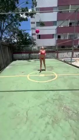 The video shows a person in a red bikini juggling a pink soccer ball on a green, fenced-in mini-soccer field. The field is marked with a yellow center circle and center line. A multi-story building is visible behind the field. The video cuts to a view of the ground and a pool area. Later, the camera focuses on a pink ball bouncing against a wall. A person’s foot in a flip-flop is visible in the frame.