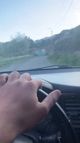The video begins with a first-person perspective of someone driving a car. The driver's hand is visible on the steering wheel, and the road ahead shows a rural setting with some houses and trees. The camera then pans to the right, showing the side mirror and the mountainous landscape outside the car window. The view transitions to a wider shot of the expansive mountain range under a bright blue sky with scattered clouds. The sun is casting shadows on the green slopes of the mountains, highlighting their rugged terrain. The video appears to capture a scenic drive through a mountainous region.