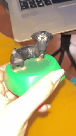 A person's hand with light-colored fingernails holds a green, bowl-shaped object. Inside the bowl rests a black dachshund-shaped toy with a blue collar. The toy is then spun rapidly within the green bowl, causing its body to rotate quickly. The toy gradually slows its spinning motion before coming to a complete stop in its original resting position. A silver laptop and a yellow surface are visible in the blurred background.