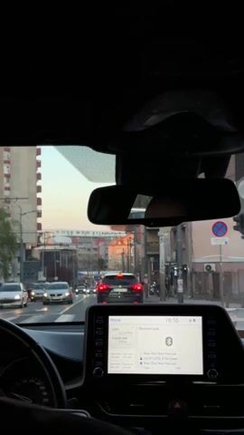The view from the interior of a car driving on a city street, at 6:56 PM, shows a traffic jam on a thoroughfare lined with buildings, and power lines overhead. The car's dashboard is in the foreground, displaying the car's infotainment system showing the audio being played. The traffic is stop-and-go. A woman walks on the sidewalk to the right.