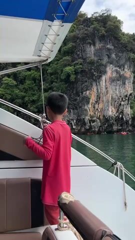 A young boy in a pink long-sleeved shirt and orange pants stands on the deck of a white boat, looking out at a rocky, tree-covered cliff face. He is holding onto a silver railing and appears to be observing the scenery. In the water below, several red kayaks are visible with people inside them. The water is a calm green color, and the sky is overcast. The boat has white and brown seating.