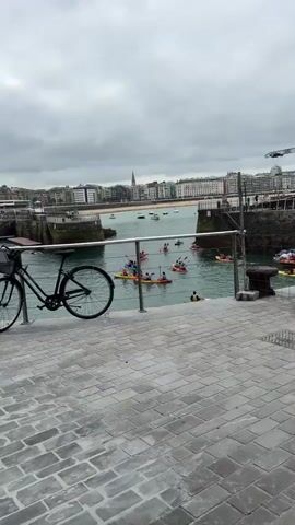 The video opens on a cloudy day with a view of a harbor. Two bicycles are parked on a cobblestone path in the foreground, with a metal railing separating the path from the water. Several kayaks are visible in the water, carrying people who appear to be paddling. In the background, buildings line the shore, and boats are docked in the harbor.

A man wearing a yellow shirt and a backpack is taking a photo with his phone. He is standing to the left of the frame, facing away from the camera. To his right, a woman in a white shirt and beige pants is leaning on the railing, looking out at the water. Another person, a man wearing sunglasses and a white t-shirt, is in the lower left corner of the frame, facing the camera and holding a phone.

The camera pans slightly to the right, showing more of the kayaks in the water and the buildings in the background. The sky remains overcast. The video then shifts to focus on the man in the yellow shirt, who is still taking pictures. The woman in beige is now closer to the center of the frame, still looking at the water. The man with sunglasses is no longer visible.

The camera continues to pan, revealing another person in the background, a woman wearing a dark top and gray pants, also looking at the water. The two bicycles remain in the foreground. The video ends with a slightly wider shot of the harbor, the kayaks, and the buildings, with the overcast sky still overhead.