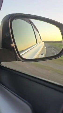The video shows a rear-view mirror of a car traveling down a road. The mirror reflects the car's passenger side, the road behind, and a sunset with a warm orange glow. Fields and trees are visible on the side of the road. The sun is low in the sky, casting long shadows. The car appears to be in motion, as indicated by the blur of the passing scenery. No people are visible in the video.