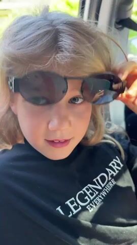 A young girl with light-colored hair and blue eyes, wearing a black t-shirt with the words "LEGENDARY EVERYWHERE," is seated indoors, possibly in a car. She has sunglasses perched on top of her head and appears to be talking. Her mouth is slightly open in what could be the middle of a word or sentence. Someone's hand is briefly visible at the top of the frame near her shoulder. The background shows the interior of a car, with the seat and some of the car frame visible. The lighting is bright, suggesting it is daytime.