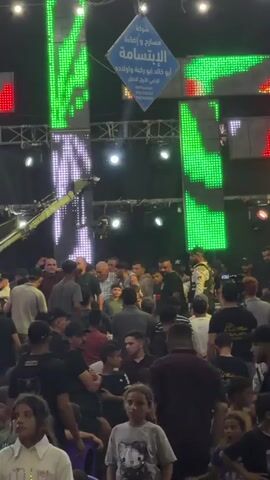 The video shows a crowded outdoor event at night. A large stage area is illuminated with bright LED lights, displaying colorful patterns and text. A banner with Arabic script hangs prominently above the stage. In front of the stage, a large crowd of people, including men, women, and children, are gathered. Some people are standing, while others are seated on plastic chairs arranged in rows. The ground appears wet, suggesting it may have rained recently. The overall atmosphere suggests a festive or celebratory occasion, possibly a performance or a public gathering.
