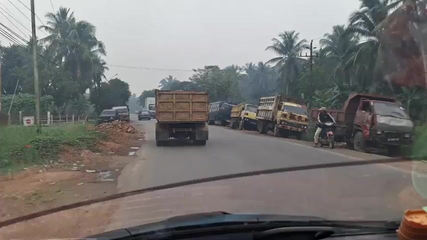 A video shows a view from inside a vehicle driving on a road lined with coconut trees. Multiple dump trucks and other vehicles are parked along the side of the road. Some trucks appear to be in a line, possibly waiting. In the foreground, the dashboard and windshield of the vehicle are visible. The sky is overcast. The road appears to be paved, with a dirt shoulder on the left side where piles of dirt and rocks are present. A white sign on a post is visible on the left side of the road. A person on a motorcycle is seen to the right of the road. The video appears to be captured during the daytime.