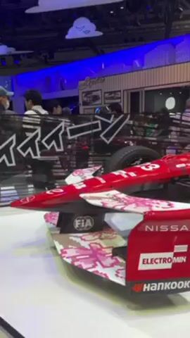 The video shows a red and white Nissan race car at a trade show. The car has several sponsor logos including Electromin and Hankook. The car is behind a small clear partition. People are walking in the background, viewing the car. The booth is decorated with skyline pictures and the name Skyline. There is a sign for Fairlady Z in the background. The car has the number 3 on the side.
