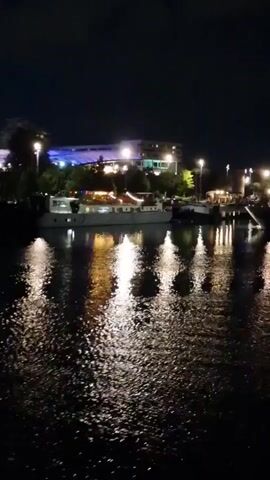 The video captures a wide shot of a waterfront scene at night. Several boats or barges are moored along the opposite bank of a body of water, which reflects numerous bright lights from the shore and the vessels. On the shore behind the boats, multiple buildings are visible, illuminated by various artificial lights, some with a cool blue hue and others with a warm yellow glow. Streetlights also dot the shoreline. The sky above is dark, indicating nighttime. The water's surface is slightly agitated, causing the reflected lights to appear as elongated, shimmering streaks. The camera remains static throughout the video.