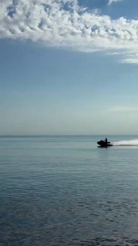 The video opens with a wide shot of a person on a jet ski, silhouetted against the bright sky, speeding across a calm blue sea and creating a long white wake. In the background, a few white clouds dot the otherwise clear blue sky.

The scene transitions to show a large, textured cliff face on the left, rising steeply from the water. The jet ski then appears from the left, approaching a red and white catamaran sailing boat with a tall mast, which is also moving through the water and leaving a wake. The jet ski drives past the catamaran.

The video then focuses on a close-up of the rugged, grey and brown rock texture of the cliff face, showing its intricate cracks and layers. The sea is visible at the bottom of the frame, reflecting the sunlight.

The footage depicts a sunny day with clear skies and calm waters in Divnomorskoe, Russia, featuring water sports and coastal scenery. No visible text is present.
