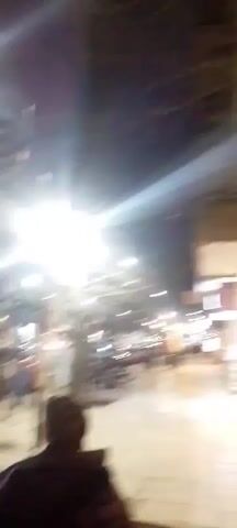 The video shows a night scene on a street with streetlights and businesses. There are cars and parked motorcycles visible. People are walking on the sidewalk. One person, likely the videographer, is in the foreground, facing away from the camera and looking towards the street. The camera pans across the street, showing more shops and streetlights. A sign with a red circle and a horizontal bar is visible on a pole.