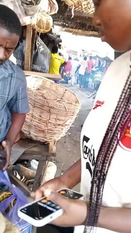 The video appears to be taken at a market or trading area.  A woman with braided hair, wearing a light-colored shirt with a red and black trim, is holding a white smartphone and using it. She's standing near a man in a blue and gray striped shirt who is examining something dark, possibly produce. Behind them is a large woven basket. Other people are visible in the background, moving around in the market setting. There is a makeshift roof overhead, constructed of straw or similar material. The environment looks like an outdoor market with vendors and customers.