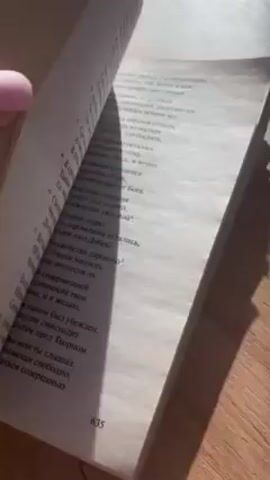 Here is a description of the video:

The video shows a close-up view of someone rapidly flipping through the pages of a thick book. The book is lying on a wooden surface that appears to be a table. The pages are thin and filled with text, likely in Russian. The light source seems to be natural sunlight. A hand with a pink fingernail is visible, holding and flipping the pages. The flipping is quick, making the text difficult to read. The page numbers are partially visible at the bottom corners, showing various numbers. Soft music is heard in the background.
