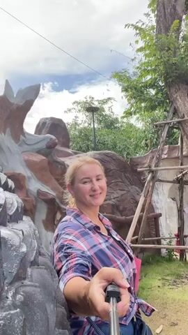 A blonde woman is taking a selfie video with a selfie stick. She is wearing a plaid shirt and is smiling. Behind her is a large statue of a person holding a staff and a pot. To the left of the statue is a large black dragon sculpture. The background consists of trees and a cloudy sky.