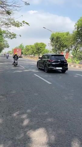This video captures a street scene in Siring, Indonesia, on a bright, sunny day with a few clouds in the sky. The asphalt road is well-maintained with clear white lane markings. The surrounding environment is lush and green, with numerous trees lining the roadside and some buildings visible in the distance. The overall atmosphere appears to be calm and ordinary, suggesting a typical day in a semi-urban or suburban area.

Several vehicles are in motion, indicating a moderate level of traffic. In the foreground, a black SUV is moving away from the camera. To its right, a white SUV is also traveling in the same direction. Further down the road, a yellow truck with a cargo bed is visible. In the distance, a red truck and a person on a motorcycle can be seen. The presence of these vehicles suggests everyday transportation and possibly commercial activity.

There are no distinct textual elements visible in the video. The time of day is clearly daytime, given the strong sunlight casting shadows. The weather appears to be pleasant and warm, typical for a sunny day. The location cues, such as the style of the vehicles and the vegetation, are consistent with a Southeast Asian setting, specifically Indonesia as indicated. The scene evokes a sense of normalcy and daily life, without any discernible emotional undertones.
