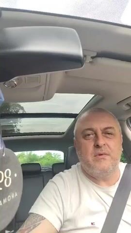 A man in a white t-shirt with short gray hair and a beard sits in the driver's seat of a car, looking directly at the camera. He points his right index finger upwards and then points it forward. Behind him, the car's back seats are visible, and a clear sky can be seen through the sunroof. The rearview mirror hangs from the ceiling. A circular air freshener labeled "AROMA" hangs from the rearview mirror.