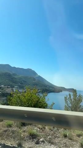 A video shows a scenic view from a moving vehicle. The camera moves past a billboard with text and images, revealing a coastal landscape. In the distance, mountains covered with green foliage rise alongside a calm blue sea. Several buildings are visible along the coastline. The foreground features a metal guardrail and some green bushes and yellow flowers. The sky is clear and bright blue.
