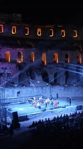 A nighttime outdoor concert is taking place within the ruins of an ancient stone structure. A brightly lit stage with blue and white spotlights is set up in the center, where a group of musicians, possibly a band, is performing. They appear to be playing string instruments and there is a keyboard or piano on stage. Several crew members are also visible on and around the stage, including one operating a video camera. The audience is seated in the foreground, facing the stage. The ancient stone walls of the structure are visible behind the stage, illuminated with purple and orange lights, highlighting the architectural details and windows. The overall scene suggests a cultural event or performance in a historical setting.