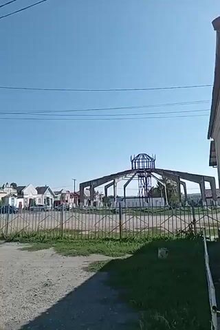 The video begins with a view of a rural street scene on a clear, sunny day. A tall lamppost stands on the left side of the frame, with power lines stretching across the sky. Several houses, some with light-colored facades and pitched roofs, are visible in the background. A chain-link fence separates the foreground from the area with the houses.

The camera then pans to the left, showing a small, light-colored building with a dark-colored roof. A dark-colored car is parked in front of it. Further down the street, other houses with light-colored walls and darker roofs are seen.

The video continues to pan right, revealing a bright yellow building with the letters "ABC" in red above its entrance. A white car is parked in front of the building, and a person is visible standing in the open doorway. The yellow building appears to be a store, indicated by the sign above the entrance which reads "МЕКТЕП МАГАЗИНИ" (School Store in Kazakh).

The camera then moves further to the right, showing a building with a red, sloped roof covered in solar panels. To the right of this building is another yellow structure, similar to the one seen earlier, with white-framed windows. Power lines and poles are visible throughout the scene, indicating the presence of infrastructure. The ground is covered with dirt and gravel. The sky is blue with a few scattered clouds. The video captures a glimpse of a simple, functional rural settlement, possibly a village or small town.