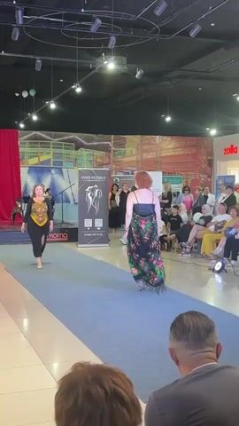 Two women are walking on a blue carpeted runway. The woman on the left is wearing a black dress with a colorful scarf around her neck. She is smiling and looking towards the audience. The woman on the right is wearing a black spaghetti strap dress with a long, floral print skirt. She is walking away from the audience and has her back to the camera. There are people sitting in the audience watching the fashion show. There is a banner behind the women with the words "MAMI MODELS" on it. There are also other banners and a red curtain in the background. The environment appears to be an indoor mall or event space.