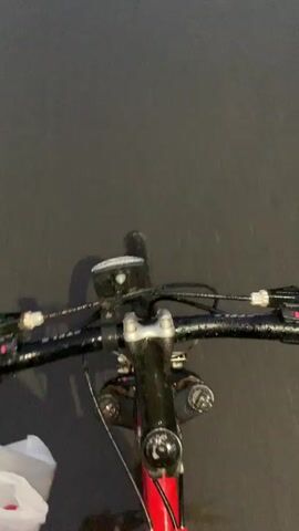 A first-person point-of-view video shows a person riding a bicycle at night on a wet road in Zvezda Snt, Russia.

The initial shot is a close-up of the bicycle's handlebars. The rider's left hand is visible, gripping the handlebar while also holding a white plastic bag with some red markings or text. A bike light is mounted on the handlebars, along with brake levers and gear shifters. The bike's frame, partially visible, is red. Faint text resembling "DESIGN" can be seen on the bike's stem.

The video then transitions to a view looking forward from the handlebars, showing the wet, dark road ahead. Reflections of distant lights are visible on the slick surface, indicating recent rain or ongoing light precipitation. The surroundings are dark, with vague shapes of trees or other vegetation along the roadside. The road appears to be a quiet, possibly residential or rural path, with no other vehicles or people visible. The activity depicted is simply cycling.