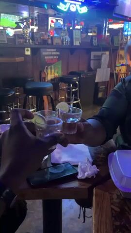 Two people are sitting at a table in a dimly lit bar, toasting with small glasses of liquor and a lime. One person is wearing a dark watch and a bracelet, and the other is wearing a dark shirt with a green camo pattern. There is a cellphone, some napkins, and a white styrofoam container on the table. Stools are positioned along a dark wooden bar lined with bottles and signs. Several televisions are visible in the background with blurred images, as well as neon signs. The music is loud and danceable.