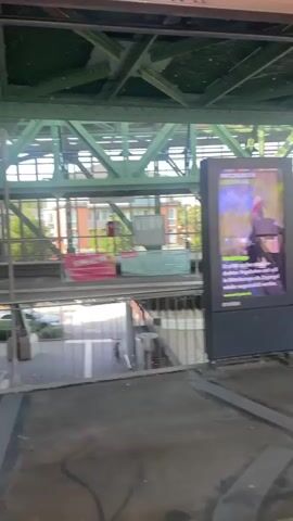 The video shows a light blue train with red accents approaching a station. The train is suspended from an overhead track and is labeled with the number 02. The train has several windows, and a person can be seen in the driver's seat. The station platform is visible, with metal railings and a sign depicting a person being prohibited from leaning. The environment appears to be urban, with buildings and trees visible in the background. The train is moving slowly and appears to be about to stop.