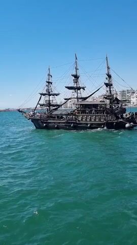 A black pirate ship with three masts sails on turquoise water under a clear blue sky. The ship is moving towards the left of the frame. On the deck, several people are visible. In the background, a city with white buildings stretches along the coastline. The water is choppy, with whitecaps.