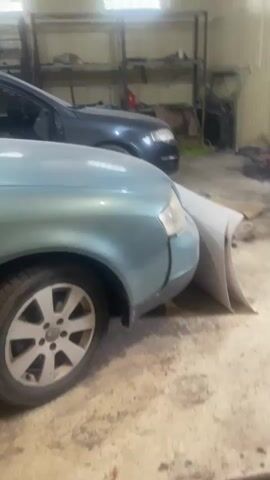 The video displays the interior of a garage or workshop. In the foreground, a light blue car with its front fender partially detached is visible, revealing its internal structure. To the right of this car, a large piece of bodywork, possibly a fender or bumper, is leaning against it. Behind the blue car, a dark gray station wagon is parked, also appearing to be in a state of disrepair or disassembly, with its doors open and interior exposed. The garage floor is concrete and appears cluttered with tools and car parts. Shelving units filled with various items are visible in the background, along with car parts hanging from the wall and stacked on the floor. The lighting suggests it is daytime, with light coming from windows or overhead fixtures. The overall impression is that of a workspace for vehicle repair or salvage.
