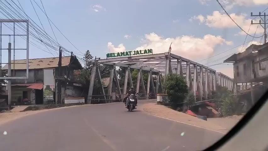 A vehicle is approaching a bridge. The bridge has a metal frame over it with the words "SELAMAT JALAN" in green lettering. There are power lines on the left. There is a building on the left and another on the right.  Vehicles are crossing the bridge in the opposite direction. There is a flag at the top right of the bridge frame. The sky is partly cloudy.