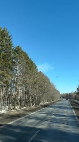 The video shows a car driving down a two-lane road on a sunny day. The sky is bright blue with a few scattered clouds. Trees line both sides of the road, with some snow still visible on the ground near the trees on the left. A vehicle is visible in the distance driving in the same direction.
