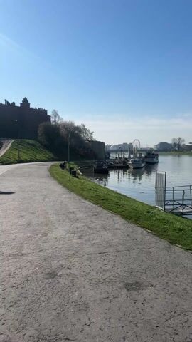 The video shows a view along a river embankment. The embankment is paved with a grey stone path bordered by a grass strip. To the left, a grassy slope leads up to a brick castle. In the background, across the river, is a Ferris wheel and other buildings. Several boats are moored to a small dock. A swan is swimming in the water. A person wearing a dark jacket and carrying a backpack walks along the path, briefly pausing near the water. The sky is clear and blue.