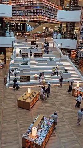 The video shows a wide shot of a large, multi-level bookstore. The dominant feature is a grand, tiered staircase with integrated seating and bookshelves. People are scattered throughout the scene, some sitting on the stairs, others walking or browsing. The bookstore's architecture is impressive, with towering shelves filled with books on multiple floors surrounding a central atrium. Decorative planetary models hang from the ceiling. The overall impression is one of a spacious and well-lit public space designed for reading and relaxation.