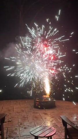 This is a video of a fireworks fountain being set off in a backyard at night. The fireworks are a mixture of white and red sparks, and they are exploding outwards from a central point. The fireworks are being set off in a fire pit, and there are two chairs and a table in the foreground. The background is dark, and there are trees visible in the distance.