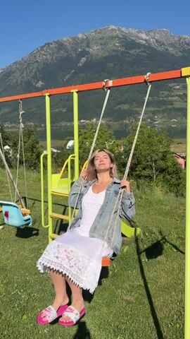A woman with blonde hair is sitting on a swing set in front of a mountain and a lake. She is wearing a white dress and a denim jacket. The swing set has a slide attached to it. The video shows her swinging back and forth. The sun is shining and the sky is blue.