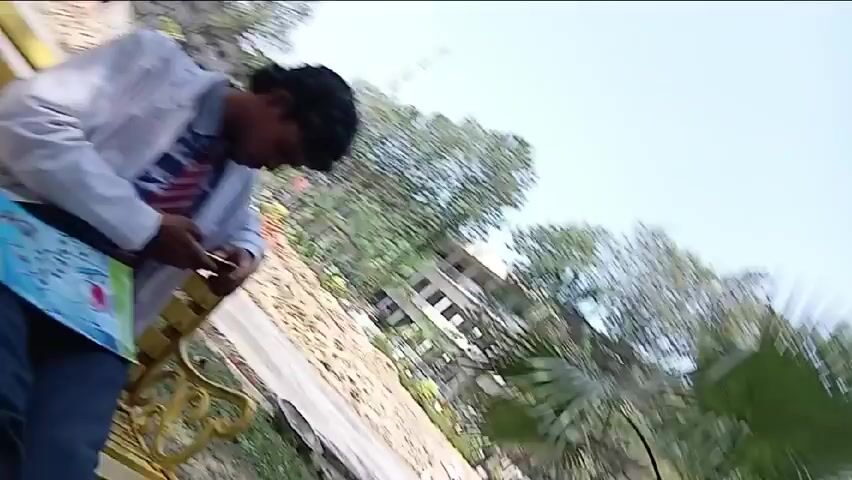 Here is a detailed description of the video:

The video is a first-person perspective shot of a person sitting on a yellow bench. The person is wearing dark blue jeans and black leather shoes. The camera focuses primarily on the person's legs and feet resting on the bench.

In the background, there is a building, trees, and other greenery, indicating an outdoor setting, possibly a park or garden area. Some people are also visible in the distance, though they are not the main focus. The lighting appears to be daytime, suggesting the video was taken in natural light.

The camera remains static for the duration of the video, with no zooming, panning, or other movements. The overall impression is of someone taking a moment to relax on a park bench and observe their surroundings.