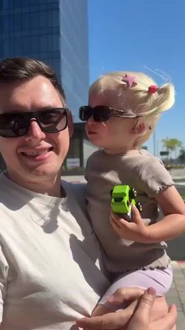 A man holds a blonde toddler in his arms. Both are wearing sunglasses. The man is on the left, smiling and laughing, and wears a white t-shirt. He makes a kissing sound and kisses the toddler on the cheek. The toddler, on the right, wears a light brown t-shirt and light purple pants, with pigtails and a star hair clip. She holds a green toy car and smiles. They are outdoors, with a modern glass building and a road visible in the background under a clear blue sky.