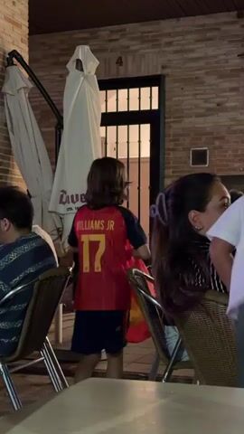 The video shows an outdoor seating area of a restaurant or bar at night. In the foreground, a child wearing a Spanish soccer jersey is visible. To the right, a woman is seated and looking towards the child. Behind her, other patrons are present, and the entrance to the establishment is visible, with people moving in and out. The setting appears to be casual and social.