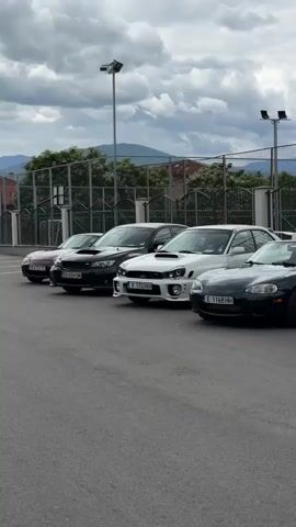 A line of cars is parked in an asphalt lot. From left to right, the cars include a black Subaru WRX, a white Subaru WRX, a dark green Mazda Miata, a blue BMW Z4, a purple Acura Integra, and a red Acura Integra. Further to the right, a white Lexus SC is partially visible. The cars are parked in front of a chain-link fence, behind which are buildings and trees. The sky is overcast with clouds.
