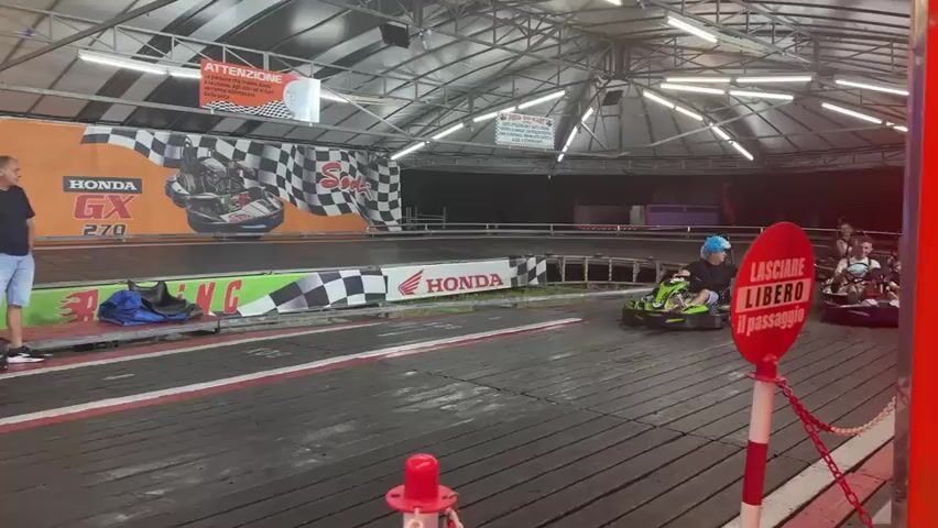 This media file captures an energetic go-karting event at an outdoor track, likely in Riccione, Italy, given the context. The scene is set under a large, open-sided canopy structure, suggesting it's either an evening event or a covered outdoor facility. The lighting appears to be artificial, with bright overhead lights illuminating the track and surrounding area.

Several individuals are present, actively participating in the go-karting. In the foreground, a person wearing a blue and white wig is driving a green and black go-kart, with their arms outstretched in a gesture that could convey excitement or surprise. Behind them, another go-kart is visible with a man and a young child inside, both appearing to be enjoying the ride. Further back on the track, more go-karts and drivers are visible, with some appearing to be lined up or waiting. A man in a blue t-shirt and shorts stands by the edge of the track, observing the activity.

The go-karts themselves are brightly colored, primarily green and black, with some featuring red accents. They are designed for racing, with low profiles and open cockpits. The track is constructed with a wooden surface and lined with red and white barriers.

Prominently displayed on the backdrop is an orange banner with black and white checkered flags, featuring the text "KARTING HONDA GX 270" and "HONDA GX 160," indicating the type of engines used in the karts and the name of the establishment. Another sign reads "ATTENZIONE" (Attention), followed by Italian text related to safety. A circular red sign with white text on a pole near the foreground says "LASCIARE LIBERO IL PASSAGGIO" (Leave the passage free).

The overall atmosphere seems lively and fun, with participants appearing engaged and possibly thrilled by the go-karting experience. The time of day is likely evening or nighttime due to the artificial lighting. The weather appears to be clear, as the canopy provides shelter but does not suggest any adverse conditions. The presence of multiple go-karts and drivers suggests a public attraction or a racing event.