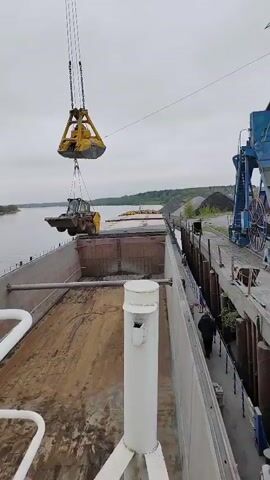 The video shows a yellow skid-steer loader being lifted by a crane and lowered into the empty cargo hold of a barge. The barge is docked next to a pier lined with material, with a river and tree-lined bank visible in the background. A figure walks along the pier alongside the barge. The day appears overcast.