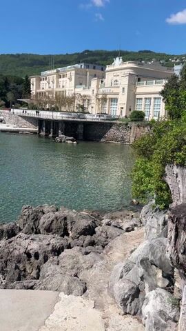 The video shows a scenic view of the Croatian coastline with calm, turquoise water. The camera pans across the water, revealing swimmers and a ladder leading into the sea. On the horizon, distant islands are visible under a clear, blue sky. The foreground features rocky formations and a concrete platform. A large, light-colored building sits along the coast, nestled against a backdrop of lush, green hills.