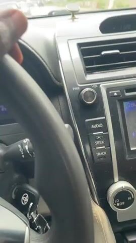 The video begins with a close-up shot of a person's hand gripping the steering wheel of a car. The dashboard is visible, showing a speedometer and other indicators. The perspective then widens to reveal the road ahead through the windshield. The car appears to be driving on a paved road in a rural or semi-rural area. Hills covered in vegetation are visible in the distance, and there are some buildings scattered along the roadside. Other vehicles can be seen on the road ahead, suggesting traffic. The sky is overcast, indicating a cloudy day. The hand on the steering wheel is dark-skinned. The video is shot from the driver's point of view.