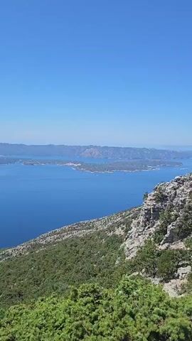 A vertical video pans across a landscape on a clear, sunny day. The foreground features green bushes and rugged, rocky terrain. A vast expanse of deep blue water, likely the sea, stretches into the distance, dotted with islands. The islands appear to be covered in vegetation and some sparse settlements. The sky above is a brilliant, cloudless blue. The camera movement is a slow, gentle pan, offering a panoramic view of the scene. There are no people or man-made structures immediately visible in the frame.