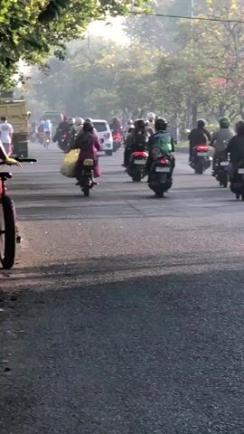 The video shows a street scene with numerous motorcycles and a few cars. The motorcycles are traveling in the same direction, creating a line of traffic. Some of the motorcycle riders are carrying items, such as a yellow bag. The environment appears to be outdoors, possibly in a city, with trees and sunlight visible. The road is paved and appears to be a multi-lane street. The lighting suggests it might be morning or late afternoon, given the long shadows. The overall atmosphere is one of a busy commute or movement of people.