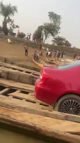The video shows a precarious situation involving cars crossing a river on a makeshift wooden bridge. Several people are gathered around a white car that appears to have trouble navigating the bridge, possibly stuck on the ramp. Another vehicle, a red car, cautiously makes its way onto the bridge. The bridge itself looks old and worn with visible gaps between the wooden planks. The river water is murky. The surrounding environment is rural with palm trees and sandy banks. Several motorcycles are parked nearby indicating that this crossing point is used by multiple modes of transportation.