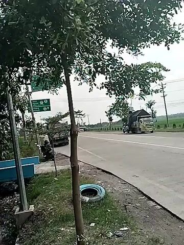 The camera captures a street view, featuring trees, a blue wall, and a passing road. A man wearing a yellow hard hat and striped shirt stands by the wall, holding a piece of equipment. A sign with "DINAS PERUM EGORO GAGENG" is partially visible in the foreground. The setting is outdoors, on a cloudy day.