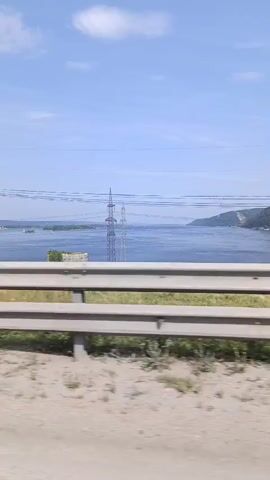The video shows a view from a moving vehicle, likely a car, traveling alongside a large body of water. In the foreground, a beige dirt road is visible, bordered by sparse green vegetation and a silver metal guardrail. Beyond the guardrail, the scene opens up to a wide blue river or lake, dotted with distant islands and lined by a green, hilly shoreline on the right. Several large, industrial-looking metal power line towers stand prominently in the middle ground, with thick cables stretching across the frame and connecting to them. The sky is clear and blue with a few wispy white clouds. The video captures the continuous movement of the landscape as the vehicle progresses.