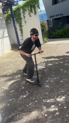 The video captures a man performing a trick on a scooter on a sunny day in Bron, France.

At the beginning, the man is seen from a side angle, standing on a black scooter. He is wearing a black beanie with "ARC'TERYX" partially visible, sunglasses, a black NFL jersey with the number 4, dark pants, and blue and yellow sneakers. He has a beard and mustache. The setting is an outdoor paved area with greenery and a building in the background.

The perspective then shifts to show the man from behind as he rides the scooter. His black NFL jersey clearly displays "CARR" and "4" on the back. He approaches a small set of stairs or a ledge. He propels himself and the scooter into the air, jumping over the obstacle. The background features a modern building with geometric white facades, green trees, and a clear blue sky with some cloud trails. A black suitcase is visible near the edge of the paved area to the left. The man lands out of frame after successfully completing the jump. The scooter deck has "ROOTS" partially visible on it.
